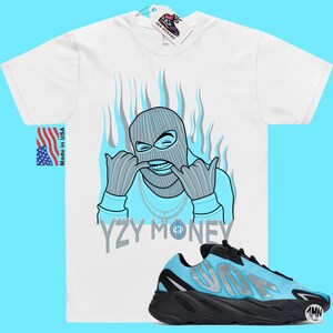 Puede incluir: Una camiseta blanca con una ilustraci&oacute;n azul y gris de una persona con una m&aacute;scara de esqu&iacute; y el texto "YZY MONEY". Debajo de la camiseta se muestra un par de zapatillas negras y azules con una franja plateada.