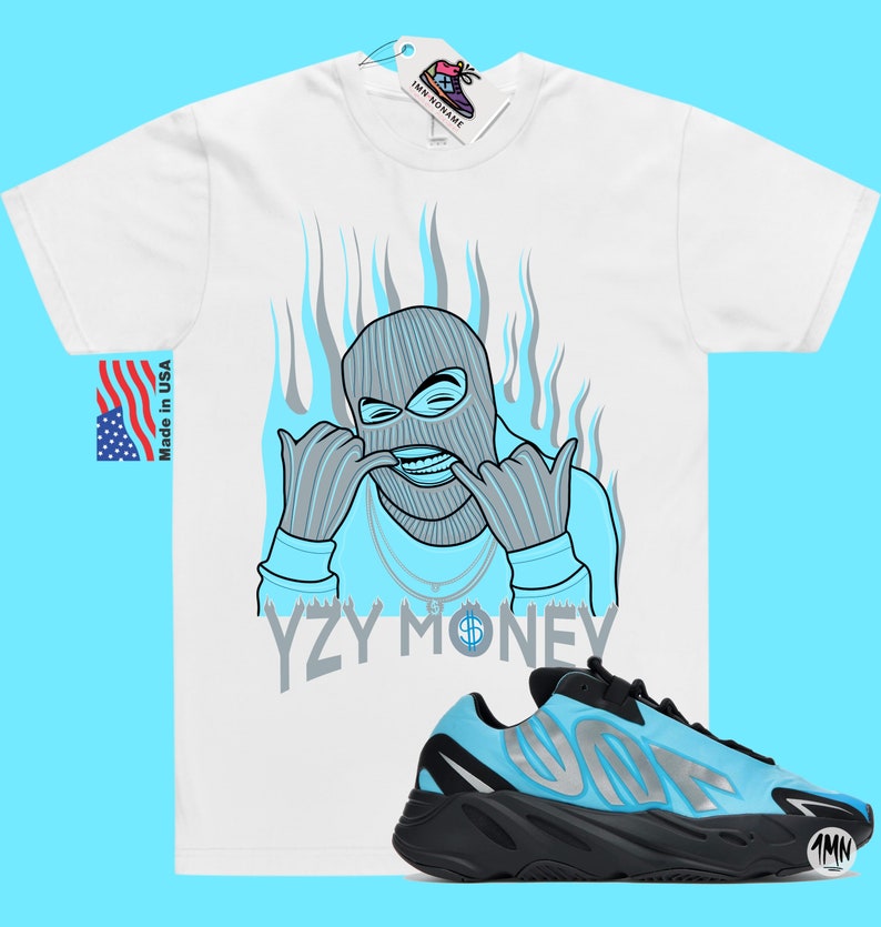Puede incluir: Una camiseta blanca con una ilustraci&oacute;n azul y gris de una persona con una m&aacute;scara de esqu&iacute; y el texto "YZY MONEY". Debajo de la camiseta se muestra un par de zapatillas negras y azules con una franja plateada.