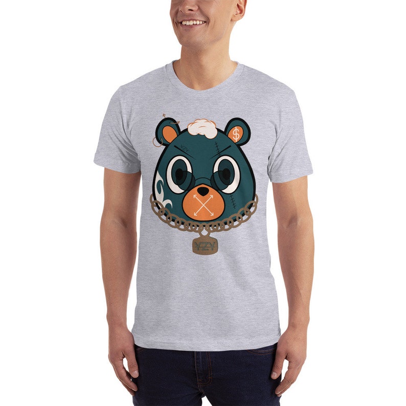 May include: A grey t-shirt featuring a cartoon bear wearing a chain with a tag that reads "YZY". The bear has a dollar sign on its head and an X over its mouth.