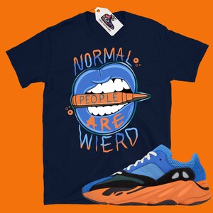 May include: A navy blue t-shirt with a graphic of a blue mouth with teeth biting a bullet that says "People are weird". The shirt is paired with orange and blue sneakers.