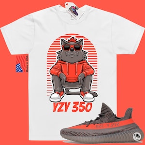Pode incluir: Camiseta branca com uma ilustra&ccedil;&atilde;o de desenho animado de um gato usando &oacute;culos de sol e um moletom vermelho. O gato est&aacute; sentado com as pernas cruzadas. O texto "YZY 350" &eacute; impresso abaixo do gato. Um par de t&ecirc;nis Yeezy 350 cinza e laranja s&atilde;o mostrados abaixo da camiseta.