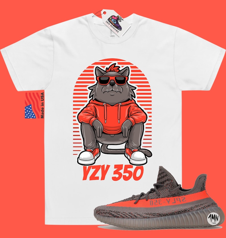Pode incluir: Camiseta branca com uma ilustra&ccedil;&atilde;o de desenho animado de um gato usando &oacute;culos de sol e um moletom vermelho. O gato est&aacute; sentado com as pernas cruzadas. O texto "YZY 350" &eacute; impresso abaixo do gato. Um par de t&ecirc;nis Yeezy 350 cinza e laranja s&atilde;o mostrados abaixo da camiseta.