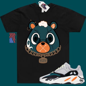 May include: Black t-shirt featuring a cartoon bear wearing a chain and a pendant that says "YZY". The bear has a dollar sign on its ear and an X over its mouth. A pair of white, green and orange trainers are shown below the t-shirt.