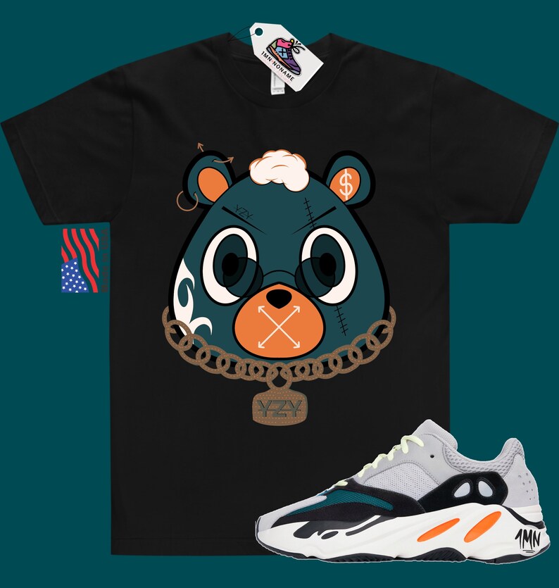 May include: Black t-shirt featuring a cartoon bear wearing a chain and a pendant that says "YZY". The bear has a dollar sign on its ear and an X over its mouth. A pair of white, green and orange trainers are shown below the t-shirt.