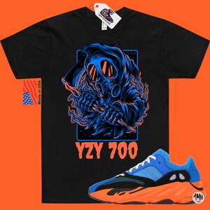 May include: Black t-shirt with a blue and black graphic of a hooded figure wearing a gas mask and holding a scythe. The text "YZY 700" is printed below the graphic. A pair of blue and orange sneakers are shown below the t-shirt.