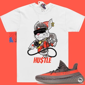 May include: White t-shirt with a cartoon rabbit wearing sunglasses, a gold chain, and a red hat. The rabbit is holding a microphone and a spray can. The text "HUSTLE" is printed below the rabbit. A pair of grey and orange sneakers are shown below the t-shirt.