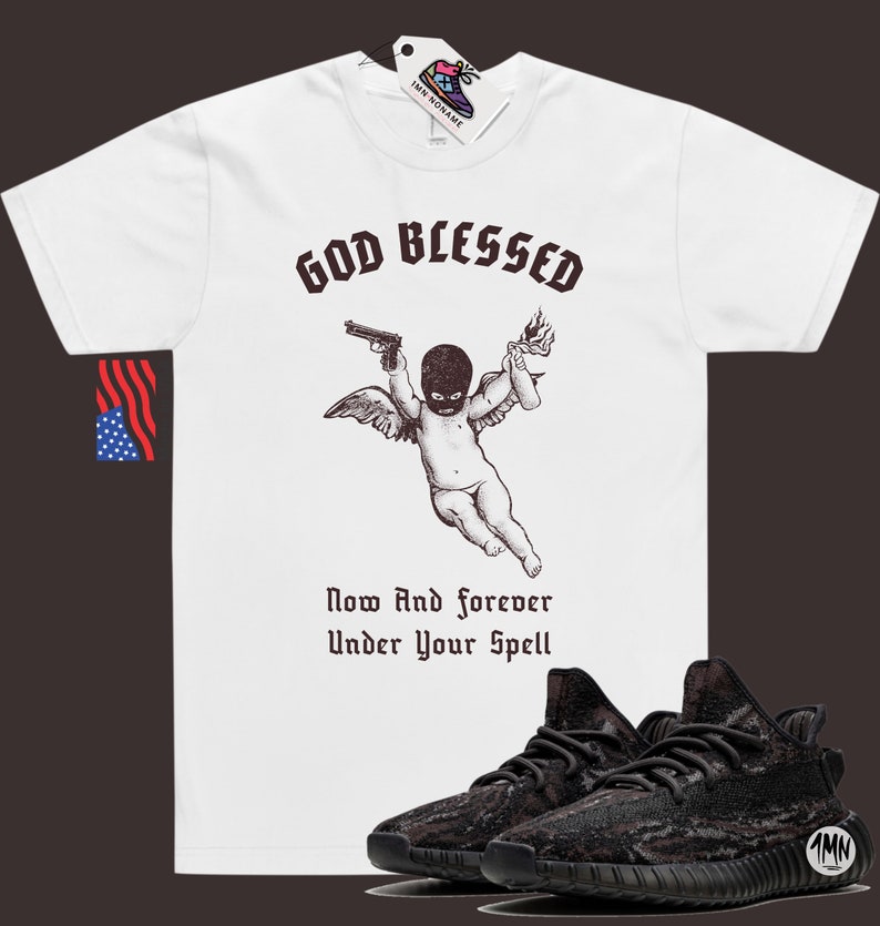 May include: White t-shirt with a graphic of a cherub wearing a ski mask holding a gun and a Molotov cocktail. The text "God Blessed" is above the cherub and "Now And forever Under Your Spell" is below. A pair of black and grey sneakers are in the bottom right corner of the image.