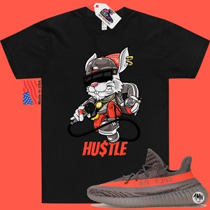May include: Black t-shirt with a cartoon rabbit wearing sunglasses and a hat, holding a microphone. The text "HUSTLE" is printed below the rabbit. A pair of grey and orange sneakers are shown below the t-shirt.