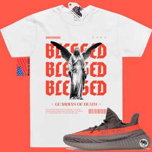 May include: White t-shirt with a red graphic of an angel with wings and the text "Blessed" repeated three times. The text "Guardian of Death" is below the graphic. A pair of gray and orange sneakers are in the bottom right corner of the image.