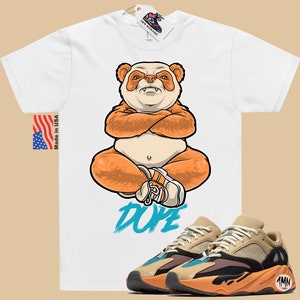 Puede incluir: Camiseta blanca con un panda de dibujos animados que lleva zapatos naranjas y la palabra "DOPE" impresa en azul. Un par de zapatillas de deporte de color beige, naranja y negro con una raya azul y el n&uacute;mero "700" en el lateral.