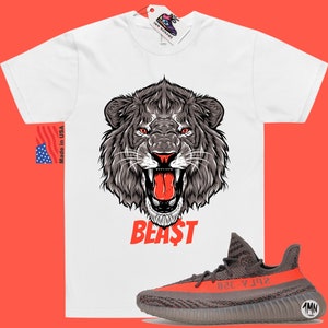 Puede incluir: Camiseta blanca con un gr&aacute;fico de la cabeza de un le&oacute;n rugiente y la palabra "BEAST" en rojo. La camiseta se combina con zapatillas grises y naranjas con el texto "YZY" en el lateral.