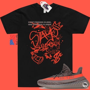 Puede incluir: Camiseta negra con estampado rojo de estilo graffiti que dice "Keep friends close, and enemies closer" y "Stay dope". Un par de zapatillas grises y naranjas con el texto "Yeezy" en el lateral.