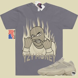 May include: A gray t-shirt with a cartoon illustration of a person wearing a ski mask and a gold chain. The text "YZY MONEY" is printed below the illustration. A pair of tan Yeezy 500 sneakers are shown in the bottom right corner of the image.