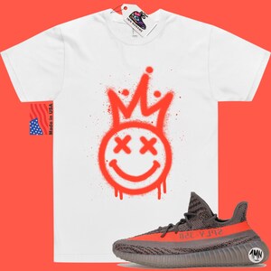 May include: White t-shirt featuring a red spray paint design of a smiley face with a crown on top. The shirt is paired with grey and orange Yeezy trainers.
