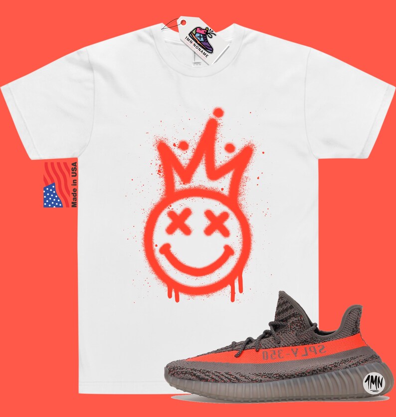 May include: White t-shirt featuring a red spray paint design of a smiley face with a crown on top. The shirt is paired with grey and orange Yeezy trainers.