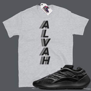 Puede incluir: Una camiseta gris con la palabra "ALVAH" impresa verticalmente en rayas negras y grises. Una zapatilla negra y gris se muestra debajo de la camiseta.