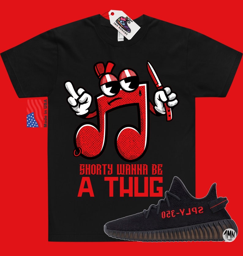 Puede incluir: Camiseta negra con un personaje de nota musical roja sosteniendo un cuchillo y el texto "Shorty Wanna Be A Thug". Debajo de la camiseta se muestra un par de zapatillas Yeezy Boost 350 V2 negras con detalles rojos.