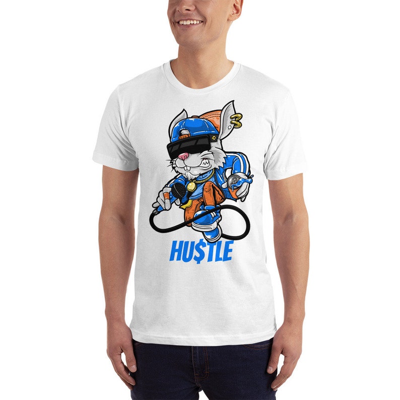 Puede incluir: Camiseta blanca con un conejo de dibujos animados que lleva un sombrero azul, gafas de sol y una cadena de oro. El conejo sostiene un micr&oacute;fono y una lata de aerosol. El texto "HUSTLE" est&aacute; impreso debajo del conejo.