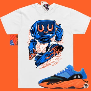 May include: White t-shirt with a cartoon illustration of a blue robot holding a pizza and riding a blue and orange skateboard. The robot has a blue television screen for a head with a cartoon face. The t-shirt is paired with orange and blue sneakers with a black sole.