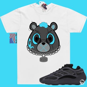 May include: A white t-shirt featuring a black and blue cartoon bear wearing a chain with a YZY pendant. The bear has a blue bandana and a stitched-up face. The t-shirt is paired with black Yeezy 700 trainers.