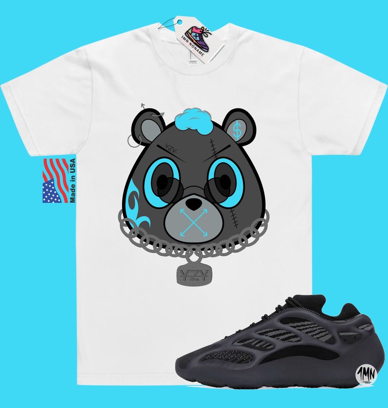 May include: A white t-shirt featuring a black and blue cartoon bear wearing a chain with a YZY pendant. The bear has a blue bandana and a stitched-up face. The t-shirt is paired with black Yeezy 700 trainers.