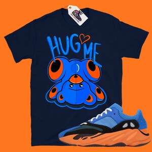 Puede incluir: Una camiseta azul marino con una criatura azul de dibujos animados con ojos naranjas y el texto "HUG ME" en naranja y blanco. Un par de zapatillas azules y naranjas se encuentran en la esquina inferior derecha de la imagen.