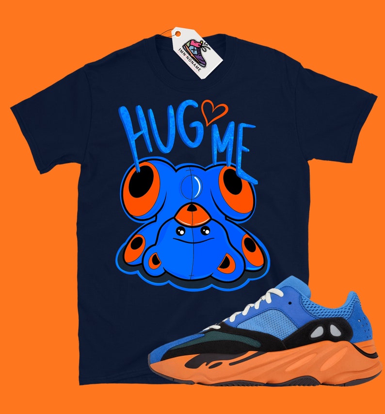 Puede incluir: Una camiseta azul marino con una criatura azul de dibujos animados con ojos naranjas y el texto "HUG ME" en naranja y blanco. Un par de zapatillas azules y naranjas se encuentran en la esquina inferior derecha de la imagen.