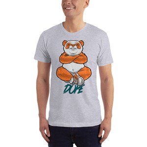 Pu&ograve; includere: Una maglietta grigia con un'illustrazione di un panda arancione in stile cartone animato seduto con le braccia incrociate. Il panda indossa scarpe da ginnastica bianche. Il testo "DUDE" &egrave; scritto in turchese sotto il panda.