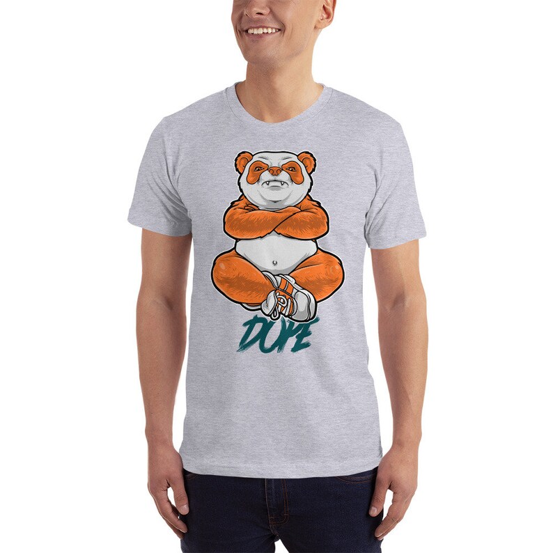 Pu&ograve; includere: Una maglietta grigia con un'illustrazione di un panda arancione in stile cartone animato seduto con le braccia incrociate. Il panda indossa scarpe da ginnastica bianche. Il testo "DUDE" &egrave; scritto in turchese sotto il panda.