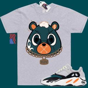 May include: A grey t-shirt featuring a cartoon bear wearing a chain and a tag that reads "YZY". The bear has a dollar sign on its ear and an X over its mouth. The t-shirt is paired with a pair of white, green, and orange trainers with the text "1MN" on the side.