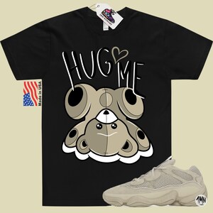Puede incluir: Camiseta negra con un oso de peluche de dibujos animados que lleva un coraz&oacute;n y el texto "HUG ME". Tambi&eacute;n se muestra un par de zapatillas beige con un dise&ntilde;o de malla.