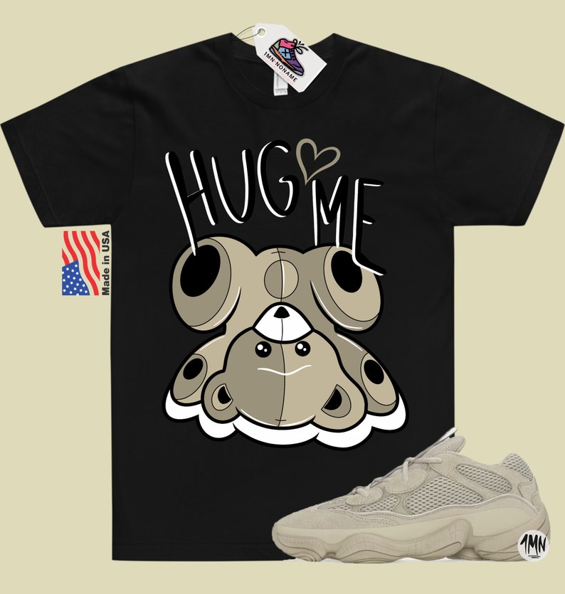 Puede incluir: Camiseta negra con un oso de peluche de dibujos animados que lleva un coraz&oacute;n y el texto "HUG ME". Tambi&eacute;n se muestra un par de zapatillas beige con un dise&ntilde;o de malla.