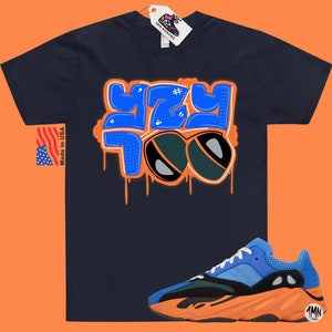 May include: A navy blue t-shirt with a blue and orange graphic that says "YZY 700" with a hashtag symbol. The t-shirt is paired with blue, orange, and black sneakers.
