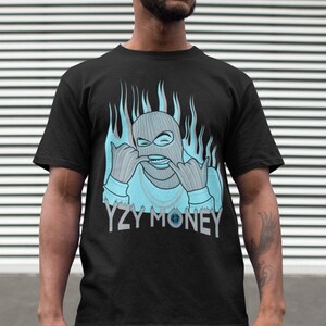 Puede incluir: Camiseta negra con un gr&aacute;fico azul de una persona con una m&aacute;scara de esqu&iacute; con llamas detr&aacute;s. El texto "YZY MONEY" est&aacute; debajo del gr&aacute;fico.