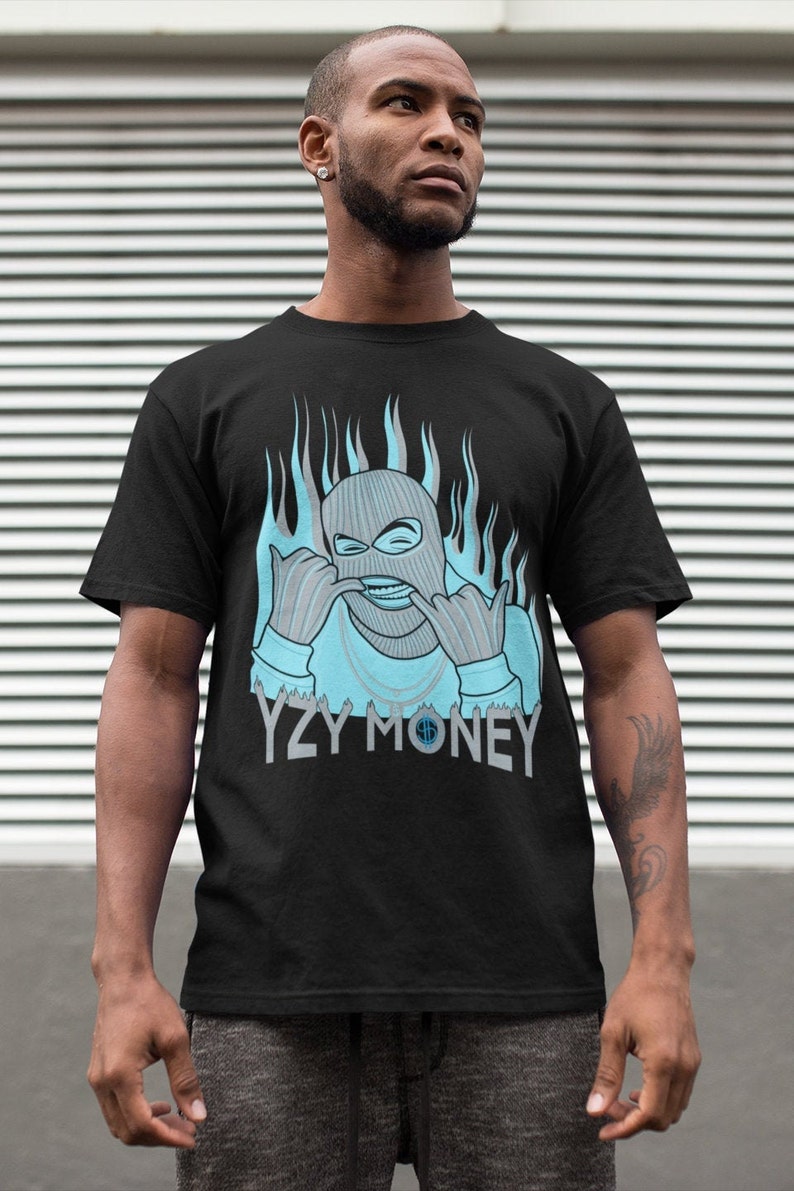 Puede incluir: Camiseta negra con un gr&aacute;fico azul de una persona con una m&aacute;scara de esqu&iacute; con llamas detr&aacute;s. El texto "YZY MONEY" est&aacute; debajo del gr&aacute;fico.