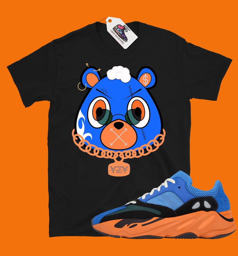 May include: Black t-shirt with a blue cartoon bear wearing a gold chain and a YZY pendant. The bear has a dollar sign on its ear and a stitched-up mouth. A pair of orange and blue Yeezy sneakers are in the bottom right corner of the image.