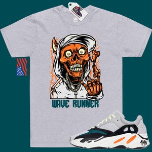 Puede incluir: Una camiseta gris con un gr&aacute;fico de una calavera con capucha y una cadena de oro. La calavera es naranja y tiene un cuerno de diablo. El texto "WAVE RUNNER" est&aacute; impreso debajo del gr&aacute;fico. Un par de zapatillas grises, negras y naranjas con el texto "Yeezy" en el lateral.