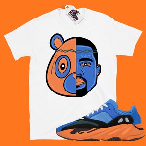 Pode incluir: Camiseta branca com um gr&aacute;fico de um urso de desenho animado e o rosto de Kanye West. O gr&aacute;fico &eacute; dividido ao meio, com o urso &agrave; esquerda e Kanye &agrave; direita. O urso &eacute; laranja e azul, e Kanye &eacute; azul. A camiseta &eacute; combinada com t&ecirc;nis laranja e azul com o texto "YZY" no lado.