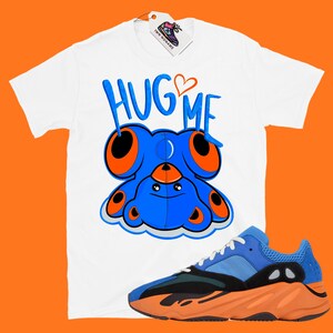 Puede incluir: Camiseta blanca con un oso de peluche azul de dibujos animados con ojos naranjas y el texto "HUG ME" en blanco y negro. La camiseta se combina con zapatillas de deporte naranja y azul.
