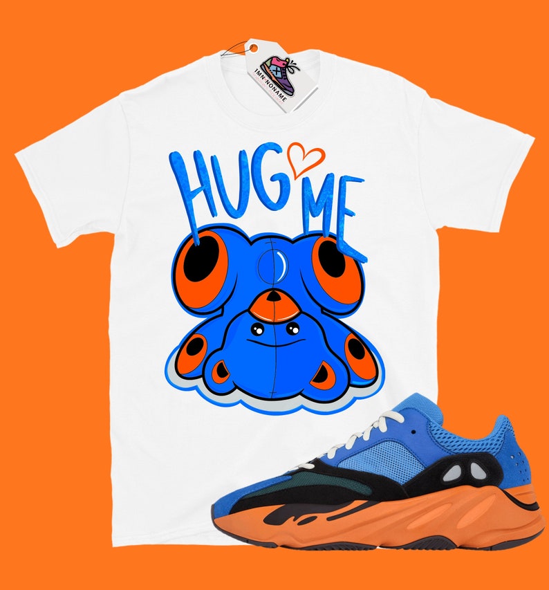 Puede incluir: Camiseta blanca con un oso de peluche azul de dibujos animados con ojos naranjas y el texto "HUG ME" en blanco y negro. La camiseta se combina con zapatillas de deporte naranja y azul.
