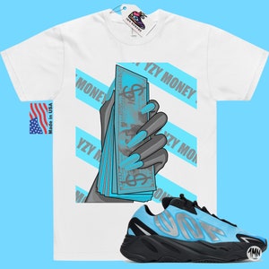 May include: White t-shirt with a graphic of a hand holding a stack of blue bills with the text "YZY MONEY" repeated across the shirt. A pair of black and blue sneakers with the text "YZY" on the side are shown below the shirt.