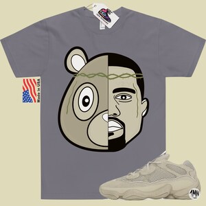 May include: A grey t-shirt with a graphic of a bear and Kanye West's face. The graphic is split down the middle, with the bear on the left and Kanye on the right. The bear is wearing a crown of thorns. The t-shirt is paired with a pair of light brown sneakers with a mesh upper and a thick sole. The sneakers have the text "1MN" on the side.