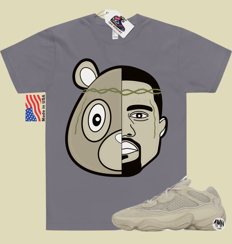 May include: A grey t-shirt with a graphic of a bear and Kanye West's face. The graphic is split down the middle, with the bear on the left and Kanye on the right. The bear is wearing a crown of thorns. The t-shirt is paired with a pair of light brown sneakers with a mesh upper and a thick sole. The sneakers have the text "1MN" on the side.