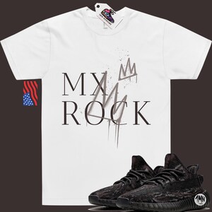 May include: White t-shirt with the text "MY ROCK" and a crown graphic. The shirt is paired with black and brown camouflage sneakers.