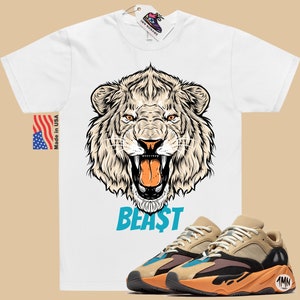 May include: White t-shirt with a detailed lion graphic and the word "BEAST" in blue. Paired with tan, black, and orange sneakers. The shirt has a "Made in USA" tag.