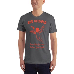 Puede incluir: Una camiseta gris oscuro con un gr&aacute;fico rojo de un querub&iacute;n sosteniendo una pistola y una antorcha. El texto "GOD BLESSED" est&aacute; por encima del querub&iacute;n y el texto "Now And Forever Under Your Spell" est&aacute; por debajo del querub&iacute;n.