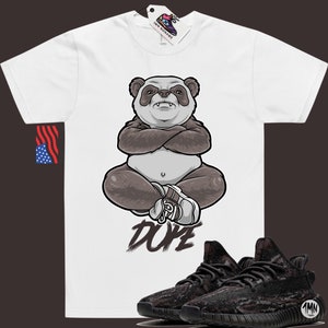 Peut inclure: T-shirt blanc avec un panda dessin anim&eacute; portant une chaussure blanche et assis en tailleur. Le texte "DOPE" est &eacute;crit sous le panda. Une paire de baskets noires et brunes &agrave; motif camouflage est repr&eacute;sent&eacute;e sous le t-shirt.