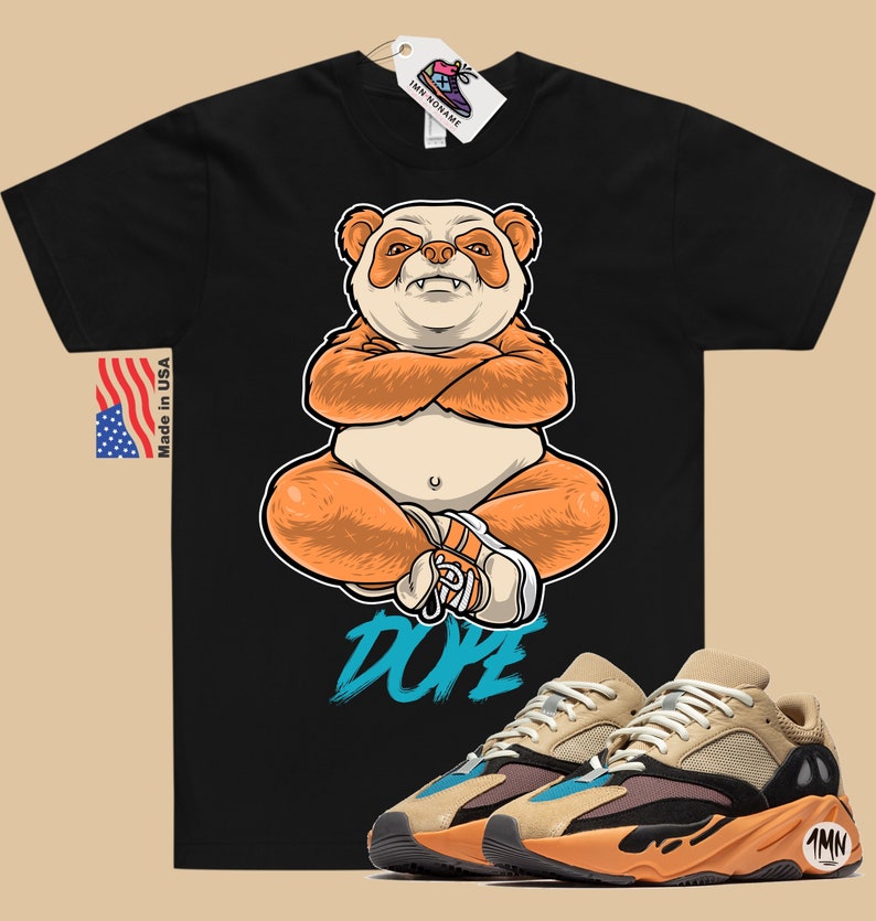 Puede incluir: Camiseta negra con un panda de dibujos animados que lleva un sombrero y est&aacute; sentado con las piernas cruzadas. La camiseta tiene la palabra "Dope" escrita en azul. Un par de zapatillas de deporte de color beige, naranja y azul con la palabra "1MN" en el lateral se muestran debajo de la camiseta.