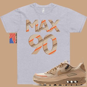 May include: A grey t-shirt with the text "MAX 90" in brown and orange stripes. The shirt is paired with a tan and orange Nike Air Max 90 shoe.