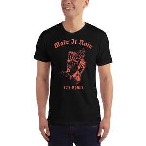 Puede incluir: Camiseta negra con un gr&aacute;fico rojo de manos sosteniendo dinero y el texto "Make It Rain" y "YZY MONEY".